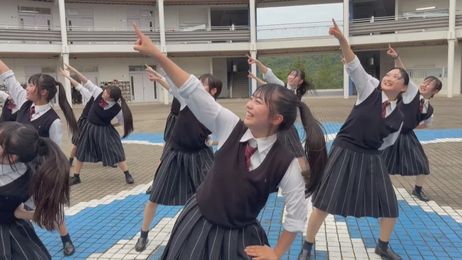 女子高生が制服姿で屋外で楽しそうにダンスのようなポーズをとっています。