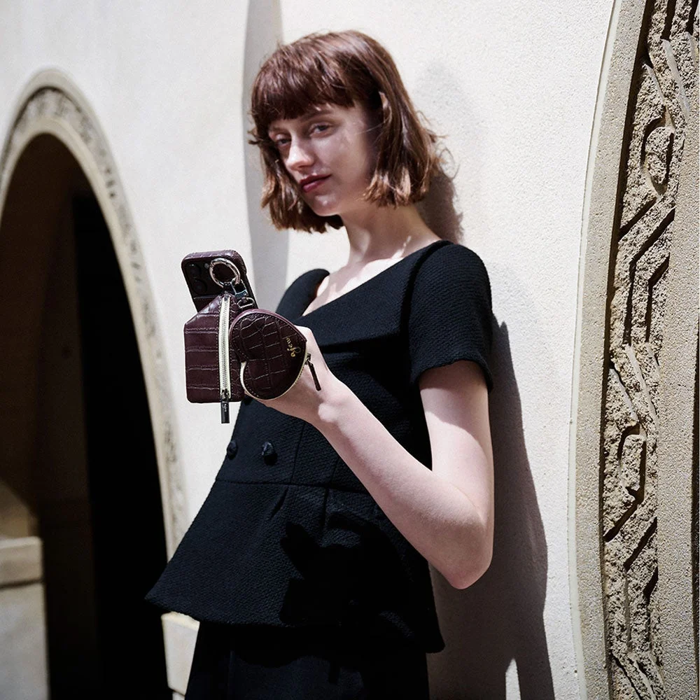 黒い服を着たショートヘアの女性が、壁にもたれかかりながら、ブラウンのスマホケースとハート型ポーチを手にしています