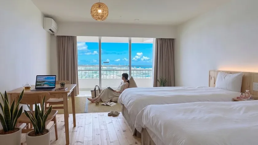海が見える明るく開放的な部屋で、女性が窓辺でリラックスして本を読んでいます。室内にはベッドやワーキングスペースもあり、快適な滞在やテレワークにも適した空間です。