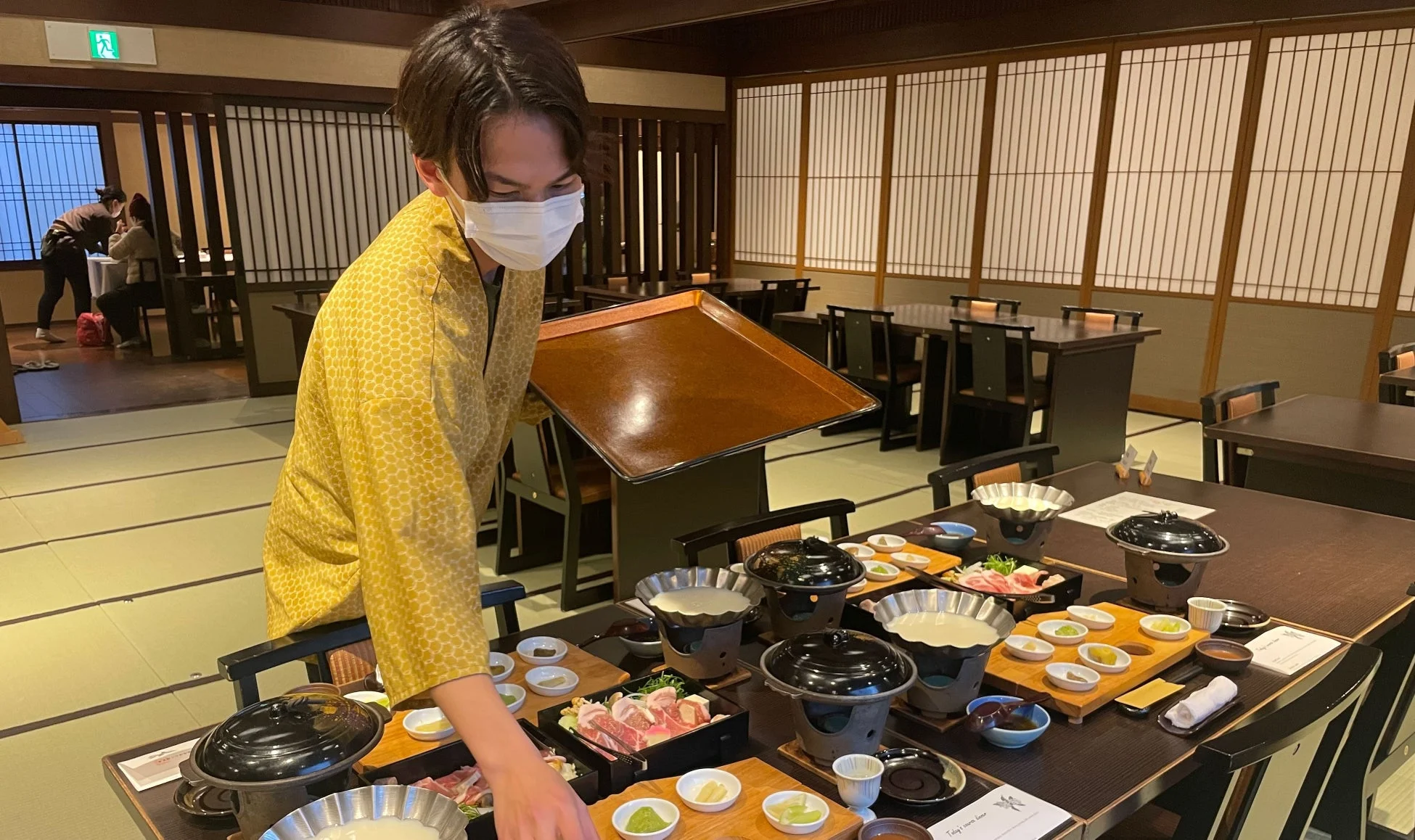 マスクと黄色の和装を身につけたスタッフが、和室のダイニングルームで個別の鍋料理の準備をしている様子。畳敷きの部屋には複数のテーブルが並び、各席には小鉢や食材が丁寧に配置され、お客様を迎える準備が整えられている。