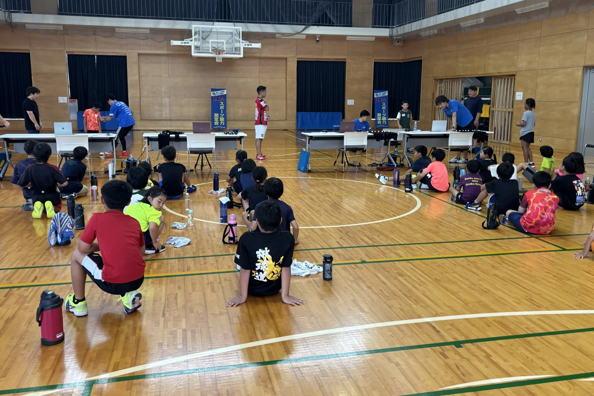 体育館で「スポーツ能力測定会」が開催されており、多くの子どもたちが床に座って説明を聞いている