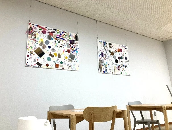 白い壁に2枚のカラフルなコラージュ作品が飾られた室内の様子。手作りのような抽象的なデザインが特徴で、下には木製のテーブルと椅子が配置されており、カフェやコミュニティスペースのような雰囲気です。