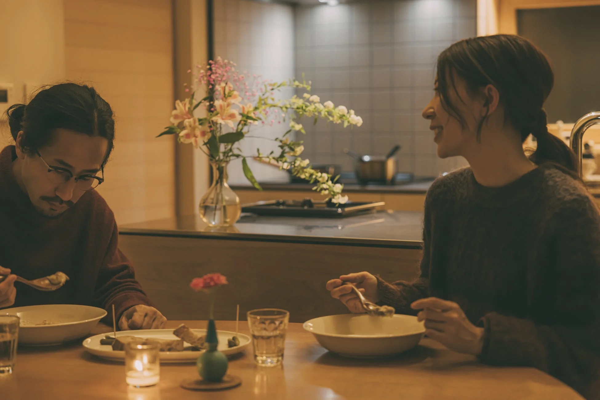 温かい照明の下、男女が食卓を囲み食事を楽しんでいる様子。会話を交わしながらリラックスした雰囲気で、花やキャンドルが飾られ、くつろいだ家庭の時間を過ごしている。