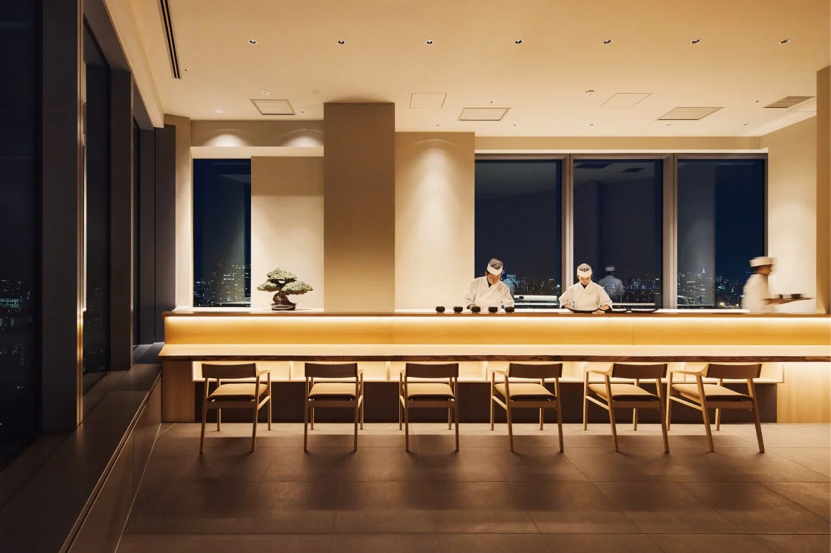 広々とした窓から夜景が見える高級感のある寿司カウンターの風景。複数の料理人が客を迎える準備をしており、モダンで洗練された和の空間が広がっています