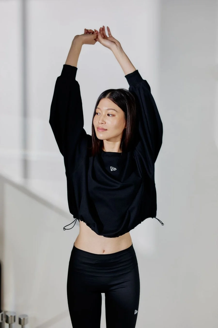 白い背景の前で、黒いスポーツウェアを着用した女性が両腕を上げてポーズをとっています