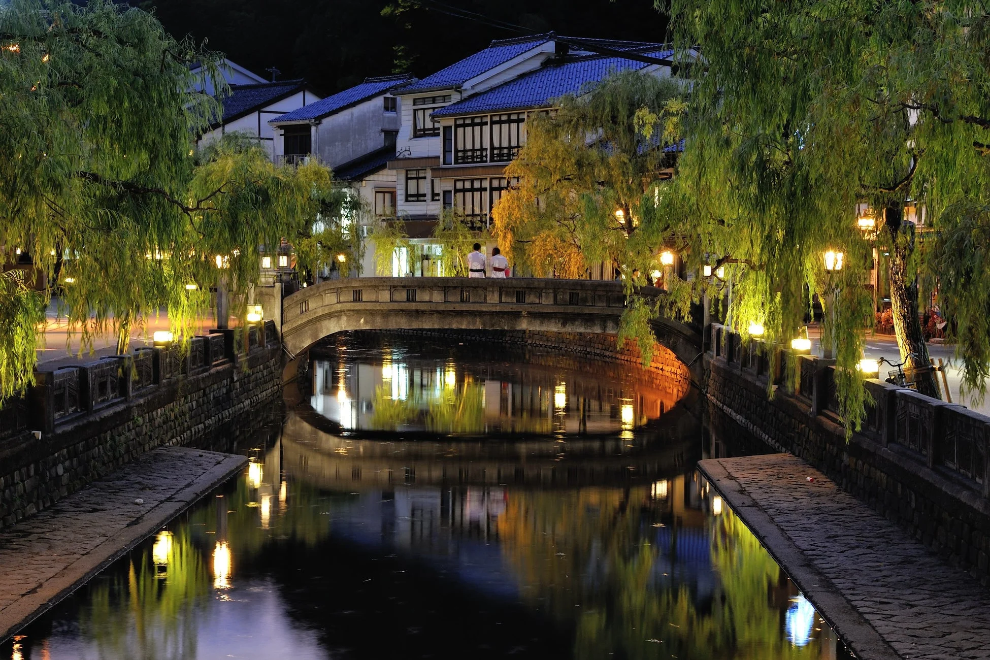 夜の日本の美しい町並みが描かれています。運河に架かる石橋の上には浴衣を着たカップルが立っており、周囲の柳の木や伝統的な家屋が提灯の光で幻想的に照らされています。水面には光が反射し、静かで風情ある風景を演出しています。