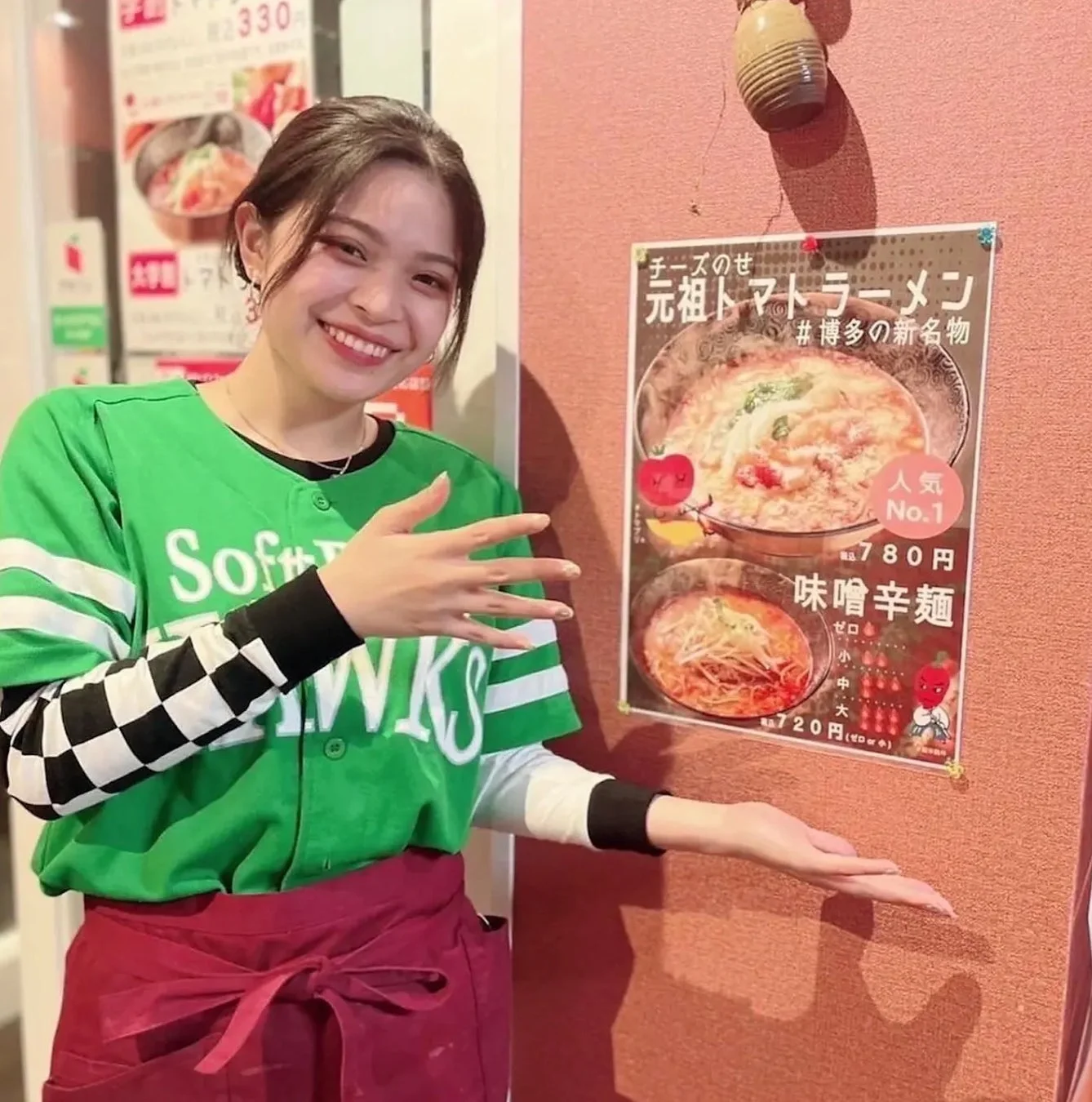 Soft Hawksユニフォームの女性が笑顔で、チーズのせ元祖トマトラーメンと味噌辛麺のメニューポスターを紹介