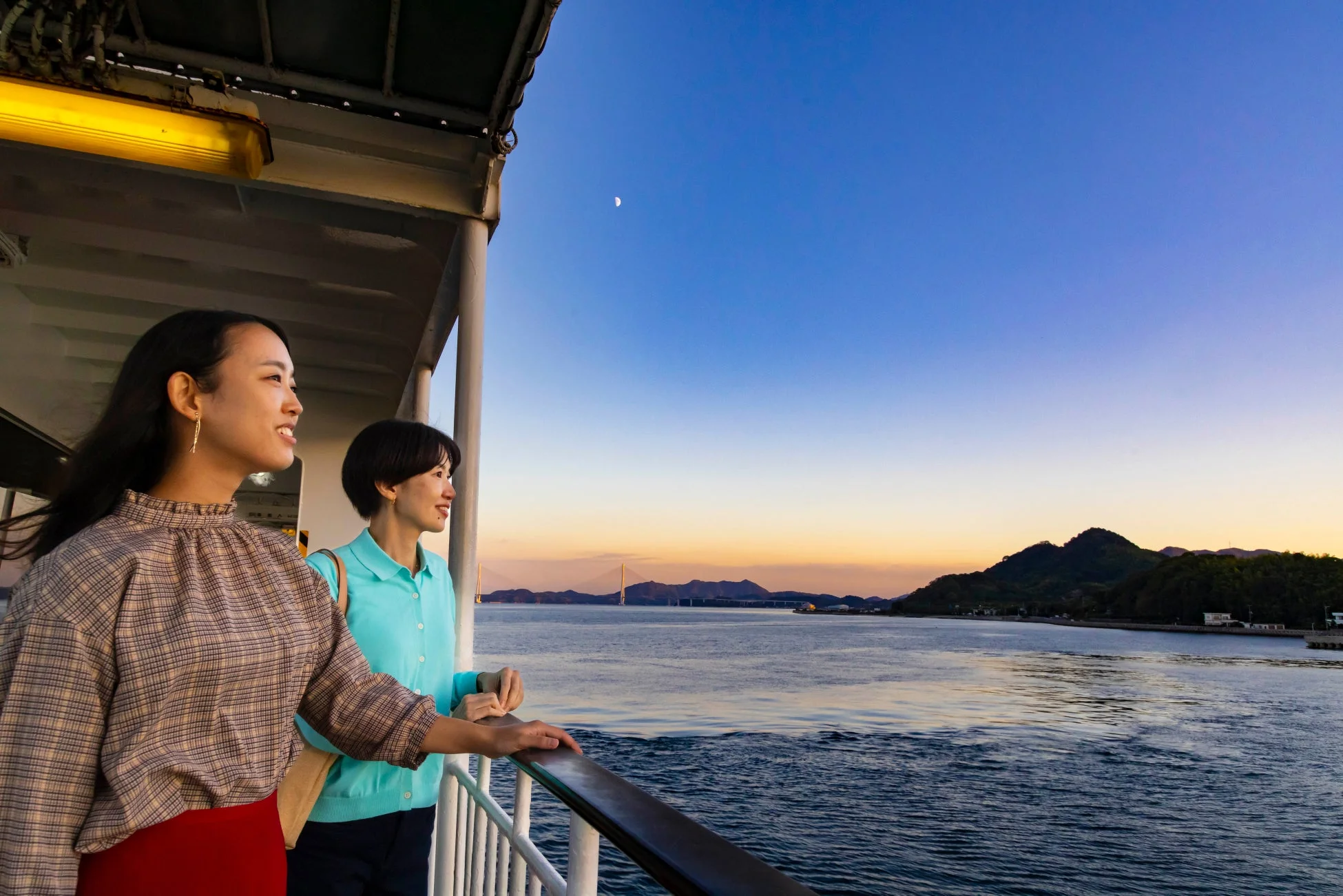フェリーのデッキから夕焼けの海と景色を眺める二人の女性。遠くには橋と山々が見え、空には三日月が浮かんでいます。楽しそうに景色を眺める姿が印象的です。