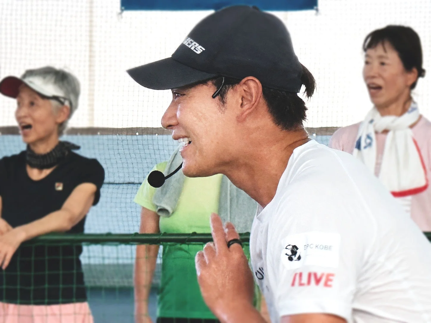 ヘッドセットを装着した男性が笑顔で話し、背景に2人の女性が写っています。スポーツ施設のような場所でのイベントやトレーニング風景で、活気ある雰囲気が伝わります。