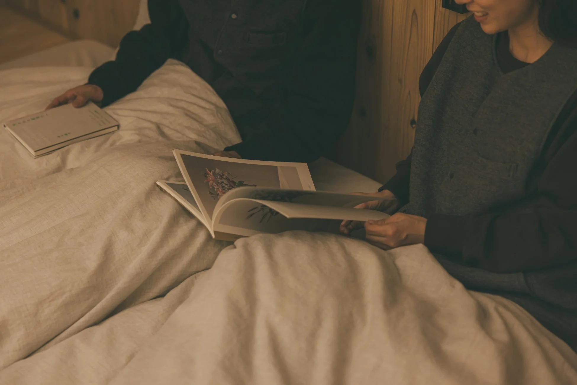 ベッドでくつろぐ二人が本を読んでいる情景です。一人が開いた雑誌を眺め、もう一人の隣には別の本が置かれています。温かく落ち着いた色調で、二人の穏やかで親密な時間が表現されています。