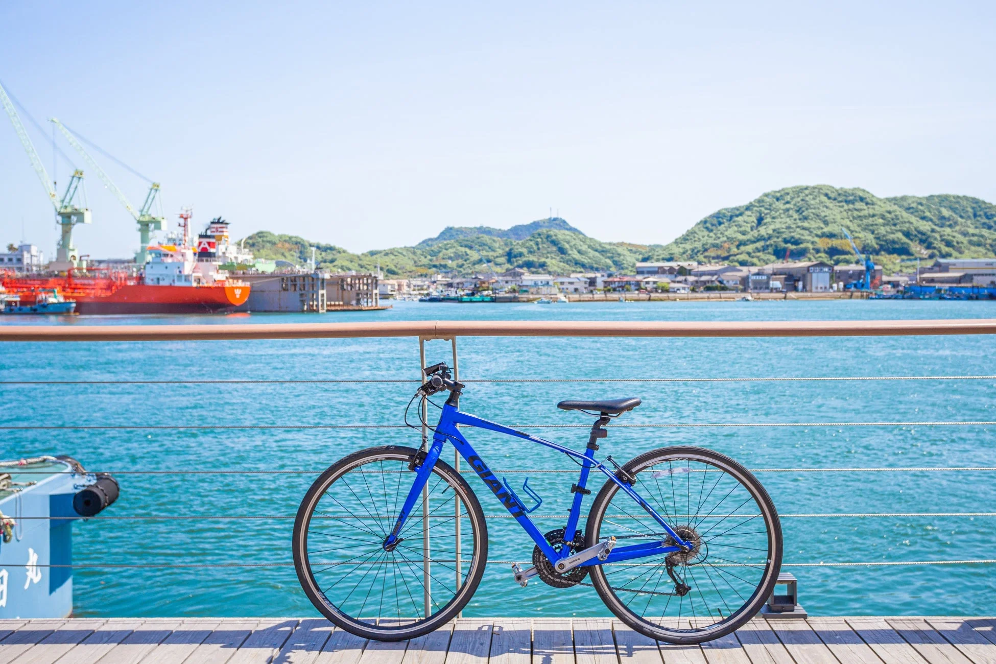 青いGIANTのクロスバイクが、晴れた日の港の木製デッキに置かれています。背景には青い海、造船所、船、そして緑豊かな山々が見えます。