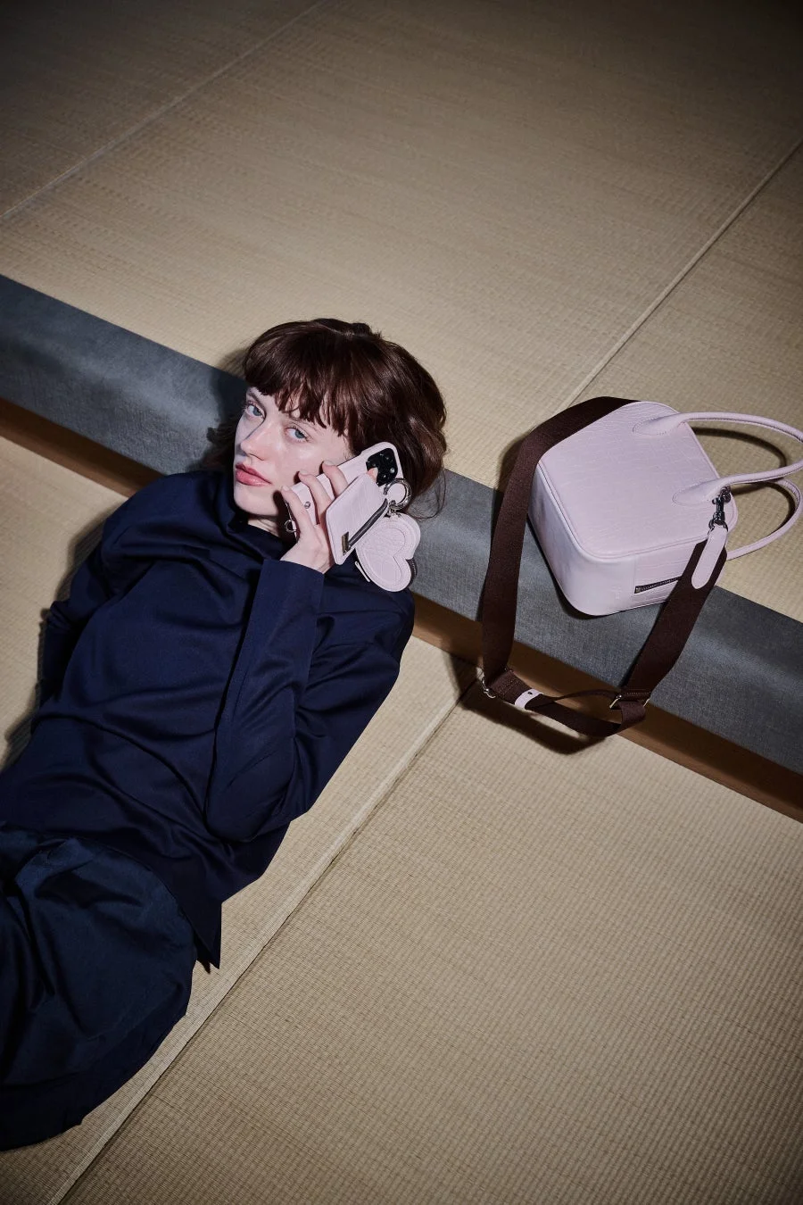 畳の上に横たわる女性が、ピンクのスマートフォンで通話しています