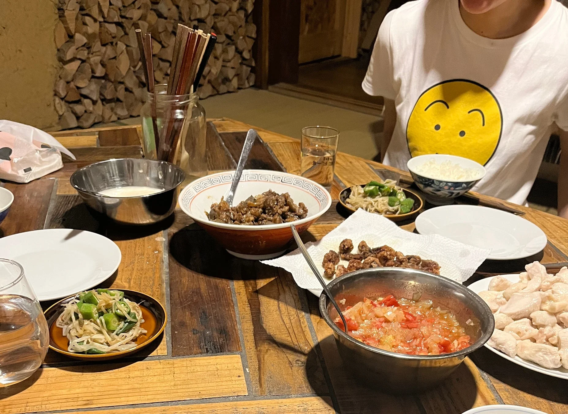 木目調の食卓に、肉料理、野菜、ご飯、鍋の具材など様々な料理が並べられています。奥には薪が積まれた壁が見え、和やかな夕食の風景を捉えています。