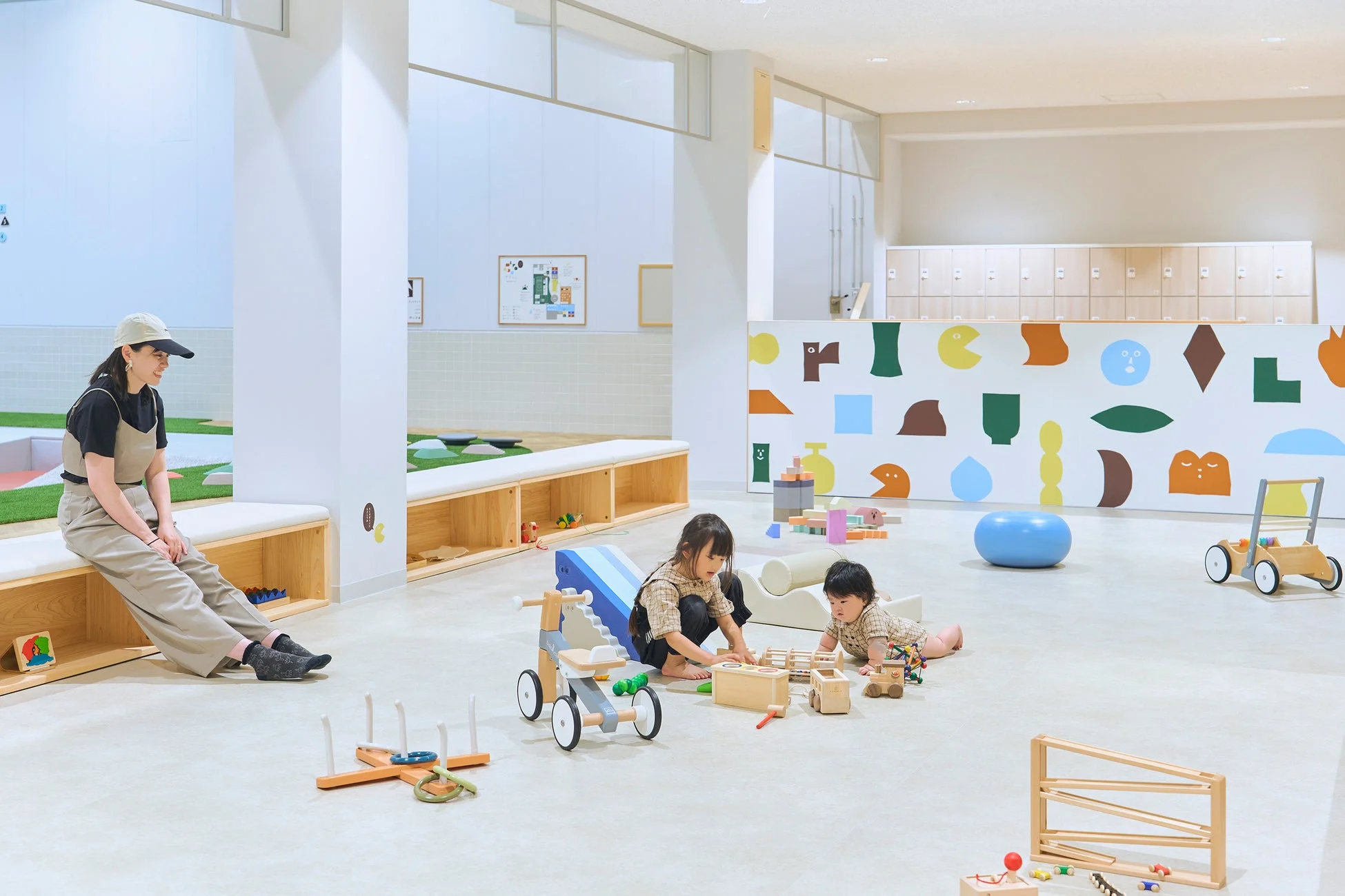 明るくモダンな室内の遊び場で、女性が見守る中、2人の子供が木のおもちゃで遊んでいます。カラフルな壁が特徴的な、清潔感のある空間です。
