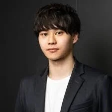 黒い背景の前で、白いTシャツの上にダークなジャケットを羽織った若い男性のポートレートです。短髪でカメラをまっすぐ見ており、落ち着いた印象を与えます。