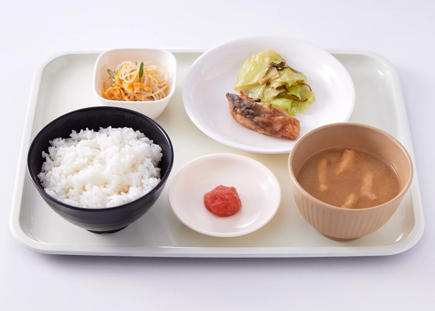 白いトレイに和食の定食が置かれています。ご飯、焼き魚と付け合わせのキャベツ、味噌汁、春雨の小鉢、明太子がバランスよく配置された健康的な食事です。