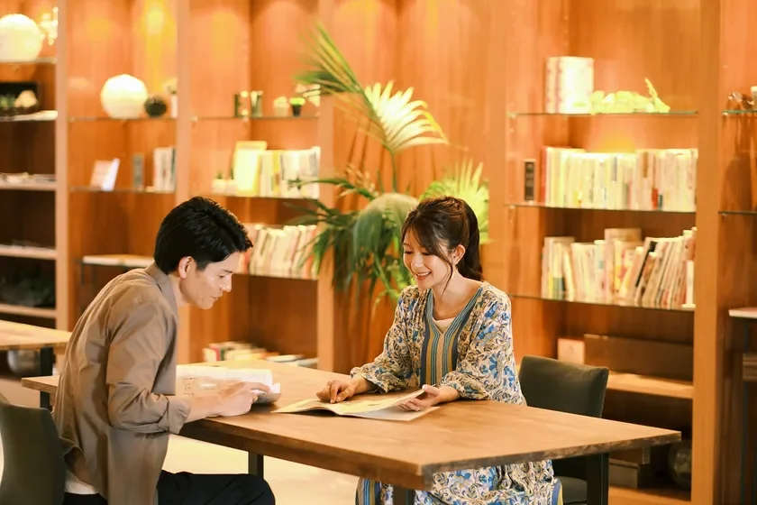 木製のテーブルで向かい合って座り、本を読んでいる男女の様子。背景には多くの本が並ぶ棚があり、落ち着いた雰囲気の空間で読書や勉強を楽しんでいる。