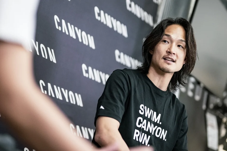 「CANYON」のロゴが並ぶ壁を背景に、黒いTシャツを着たアジア人男性