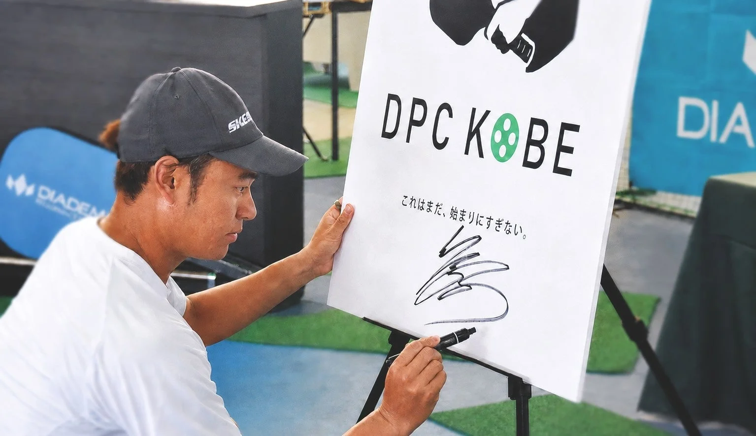 男性が「DPC KOBE」と書かれたボードにサインをしている様子。これはまだ、始まりにすぎない。というメッセージが添えられている。