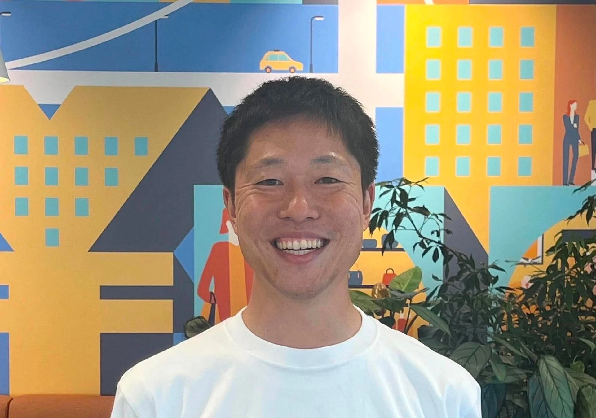 白いTシャツを着た笑顔の男性が、カラフルな抽象的な都市の壁画を背景に写っています。
