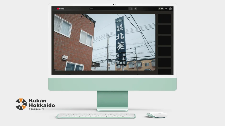 ミントグリーンのiMacの画面に、日本の街並みと「株式会社北菱」の看板が映し出されています。手前にはキーボードとマウスが置かれ、左下には「Kukan Hokkaido」のロゴが見える、モダンなデスクトップ環境です。