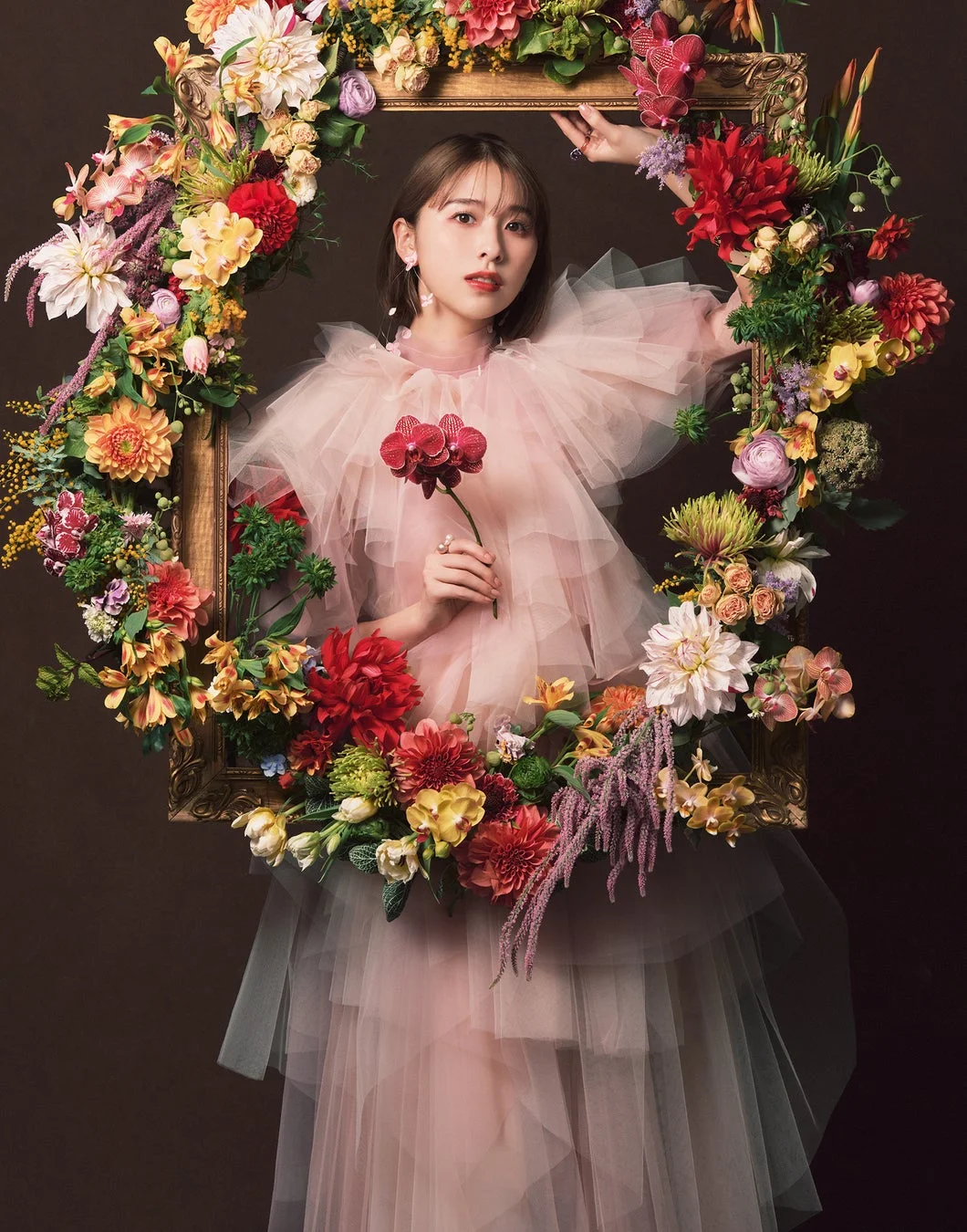 ピンクのフリルドレスを着た女性が、色とりどりの花で飾られた豪華な額縁の中に佇んでいます。