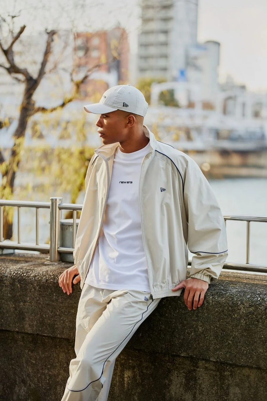 ベージュのトラックスーツと白いTシャツ、キャップを着用した男性が護岸に寄りかかっています