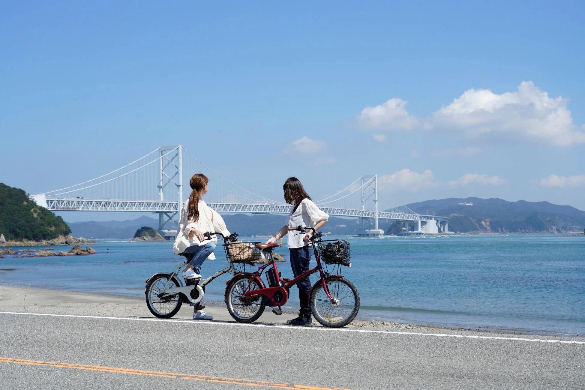 青い空と海を背景に、自転車に乗った二人の女性が橋を眺めている風景