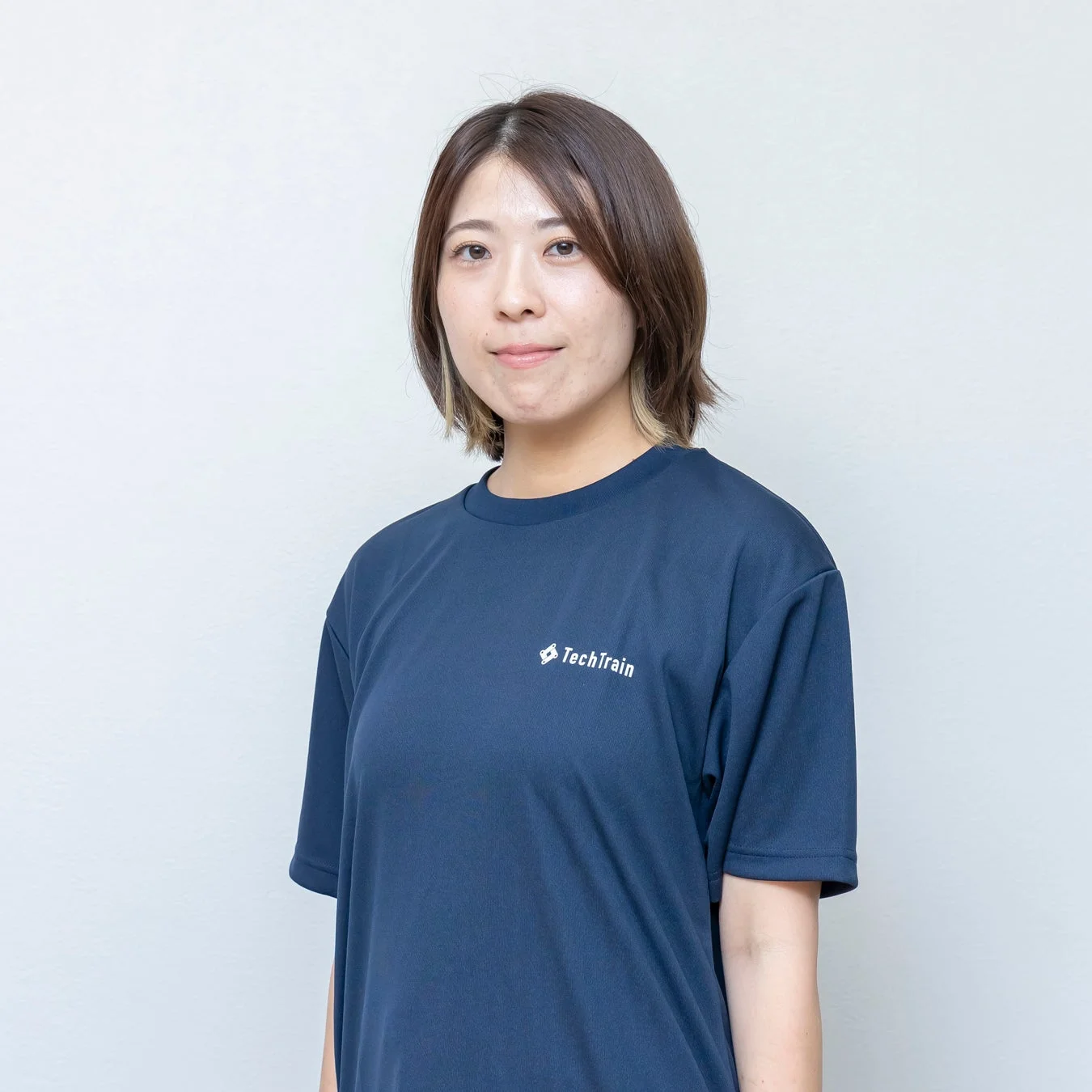 TechTrainのロゴ入りTシャツを着たショートヘアの女性が、白い壁を背景にカメラを見つめているポートレート写真です。