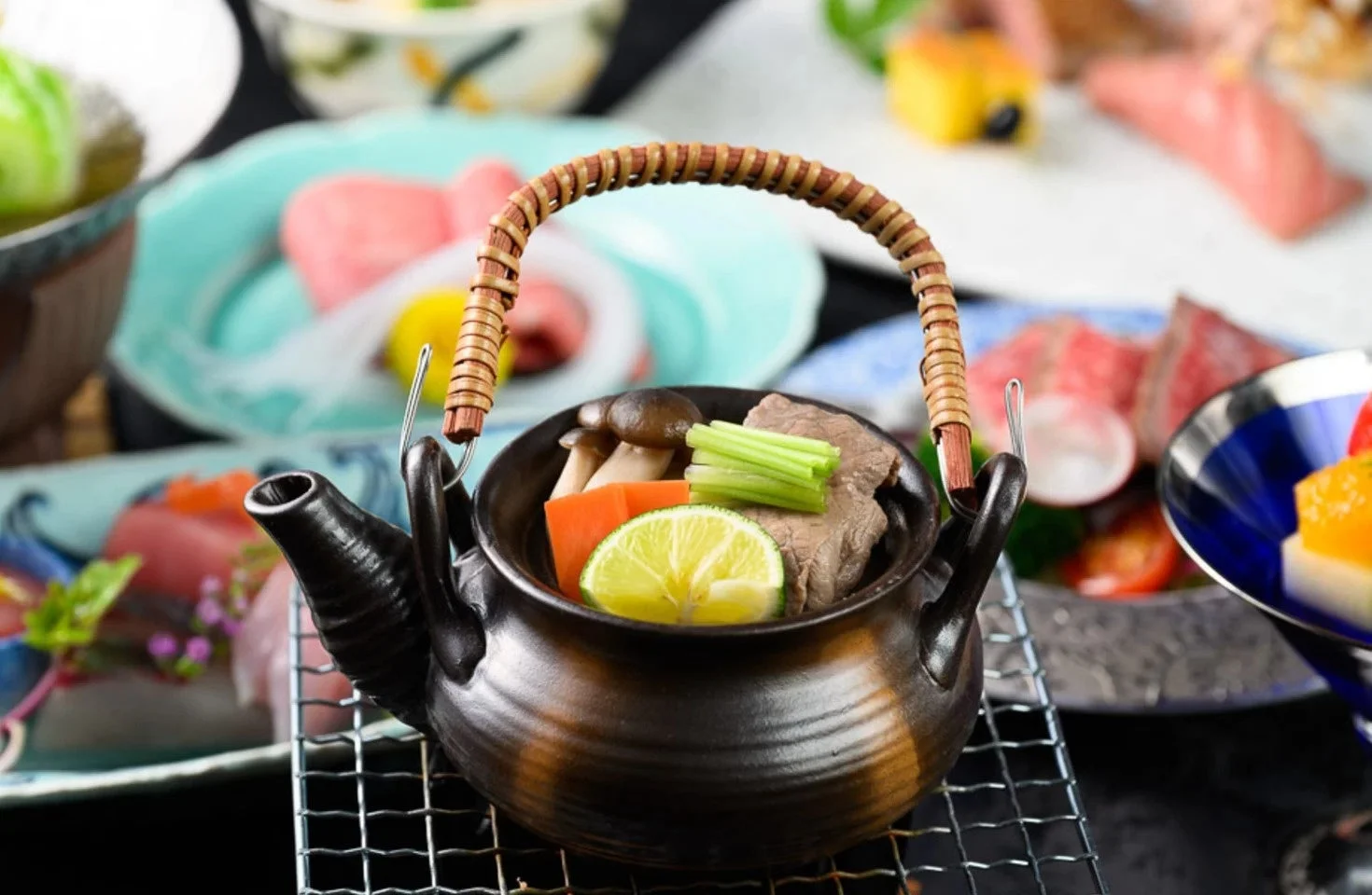 豪華な懐石料理の食卓で、手前には肉、きのこ、野菜、ライムが入った土瓶蒸しが置かれている。背景には美しい盛り付けの様々な和食が並び、伝統的で洗練された日本の美食を表現している。