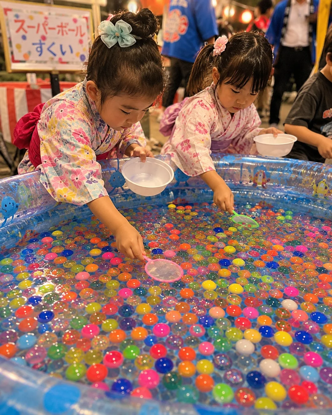 浴衣を着た2人の幼い女の子が、お祭りでスーパーボールすくいを楽しんでいる様子。