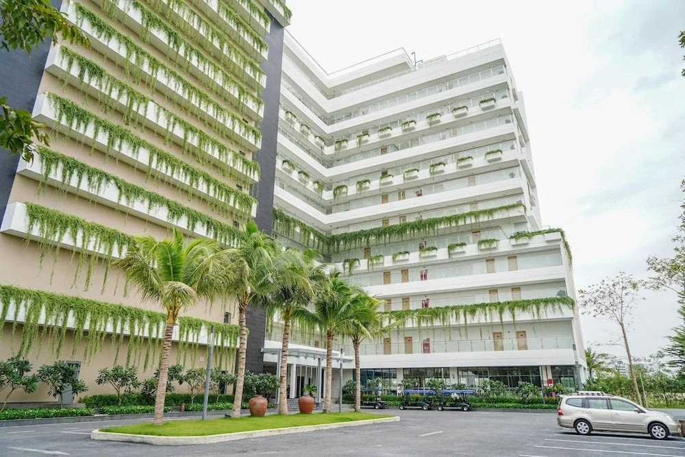 多数の植物で緑化されたモダンな建物が特徴的な風景です。建物は白とベージュを基調とし、各階のバルコニーから緑のつる植物が垂れ下がっています。手前にはヤシの木が立ち並び、駐車場には数台の車が見えます。熱帯地域のリゾートホテルかマンションのような雰囲気です。