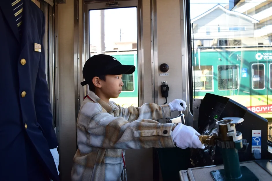 電車の運転席で、白い手袋をはめた少年が運転操作を体験している様子