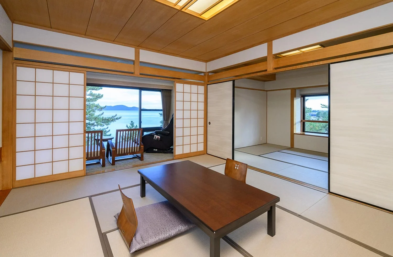 畳敷きの和室で、大きな窓からは海または湖と山々が望める絶景が広がっています。室内には座卓と座椅子、さらに窓辺にはリラックスできる椅子が配置されており、伝統的ながらも快適な空間が感じられます。