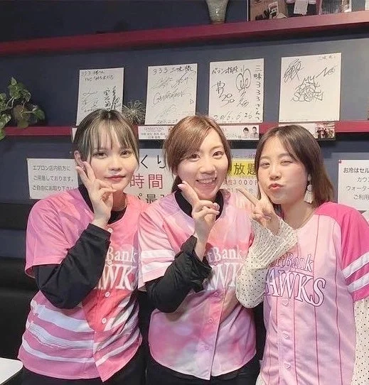 ピンクのソフトバンクホークスユニフォームを着た3人の日本人女性が笑顔やピースサインでポーズ