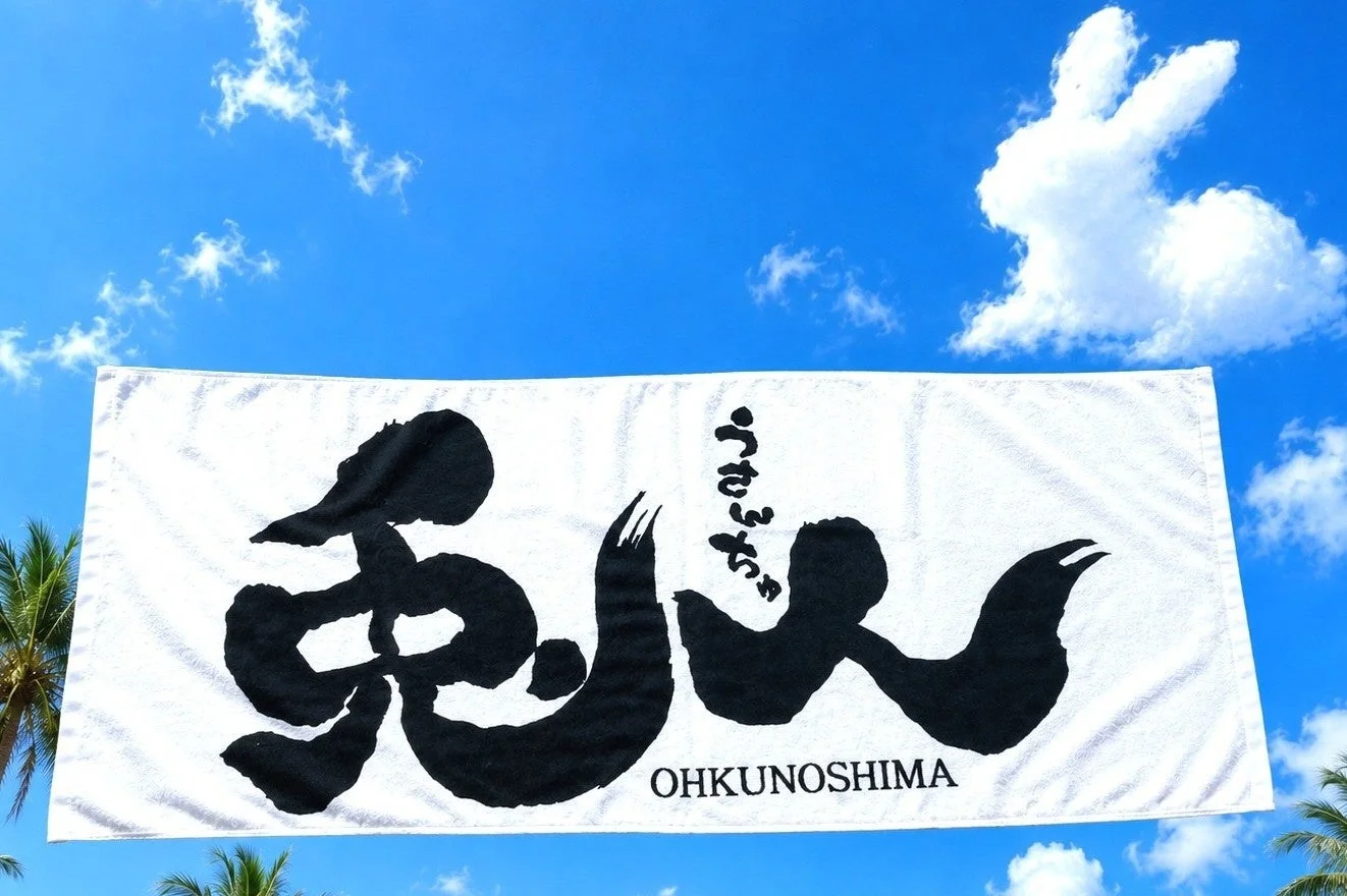 青空の下、大久野島の名前が書かれた白いタオルが掲げられています。上空にはうさぎの形をした雲が浮かび、南国らしいヤシの木も見えます。大久野島（うさぎ島）を象徴する風景です。