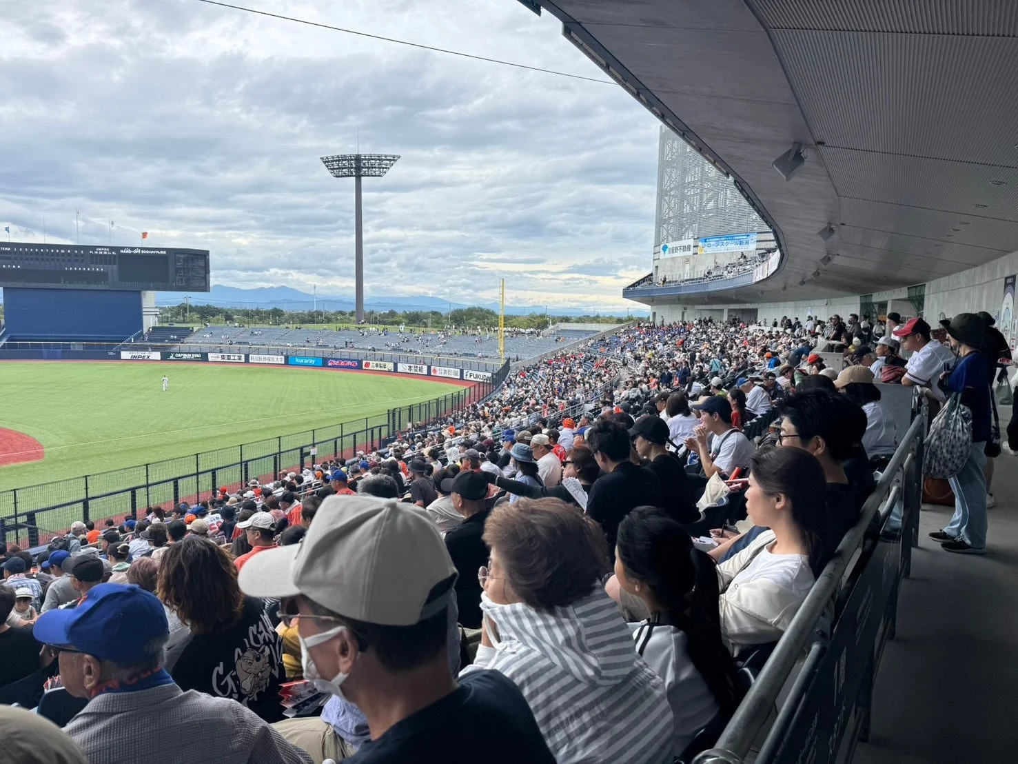 野球場で多くの観客が試合を観戦している様子。緑のグラウンドでは選手がプレーしており、大型ビジョンにはスコアが表示されている。曇り空の下、活気あるスポーツイベントが開催されている。