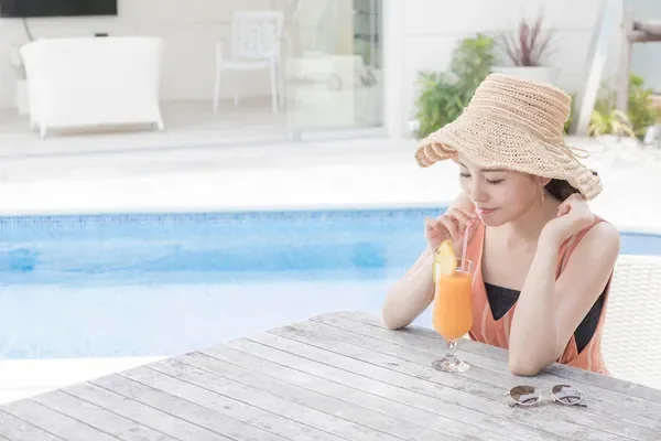 麦わら帽子をかぶった女性がプールサイドの木製テーブルでドリンクを飲んでいます