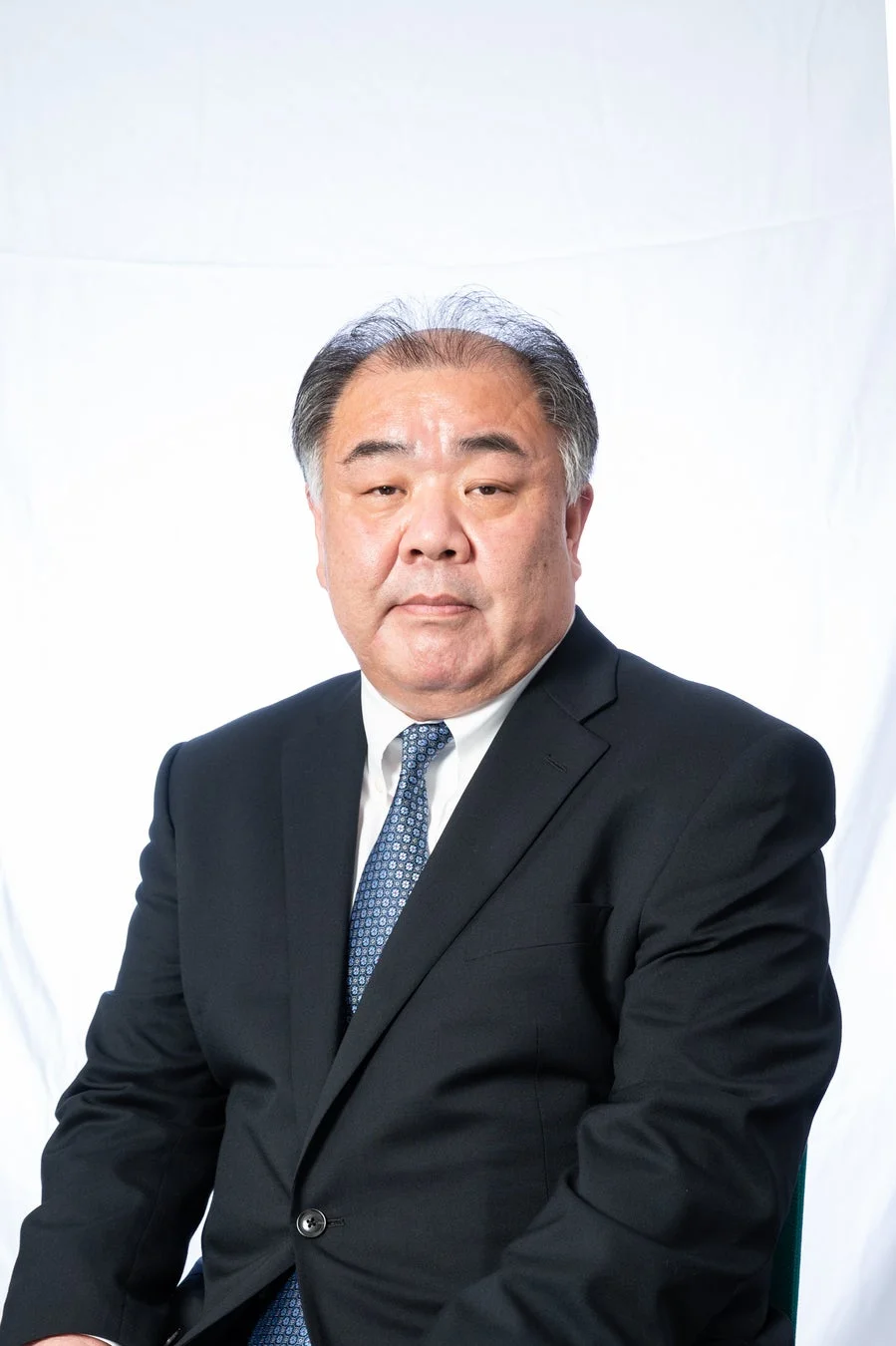 白を背景に、ネイビーのスーツと水色のネクタイを着用した中高年の男性が正面を向いている