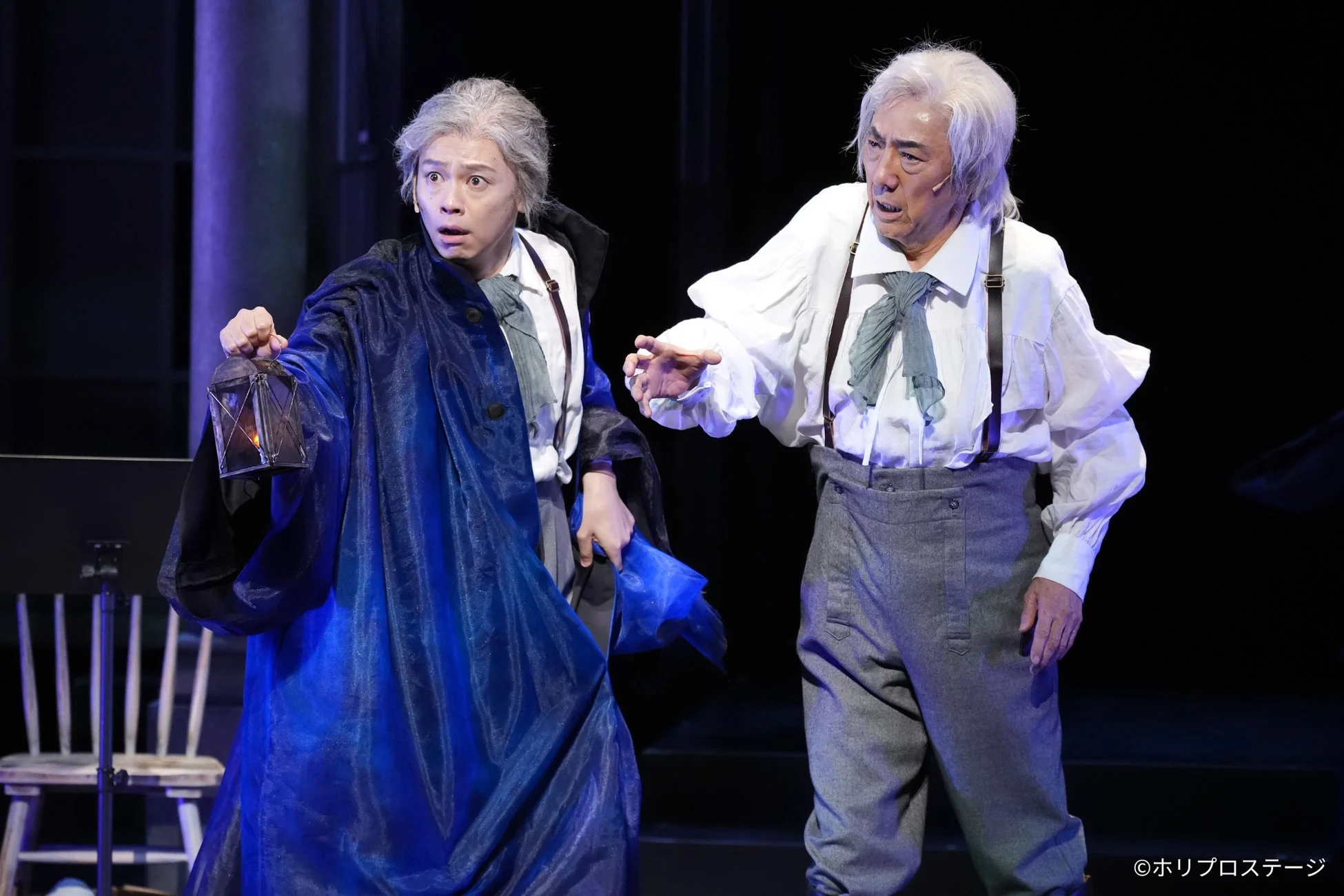 舞台上で演技をする2人の男性俳優の様子。左の男性は青いローブをまといランタンを持ち驚いた表情で、右の男性は白いシャツとサスペンダー姿で何かを訴えかけている。
