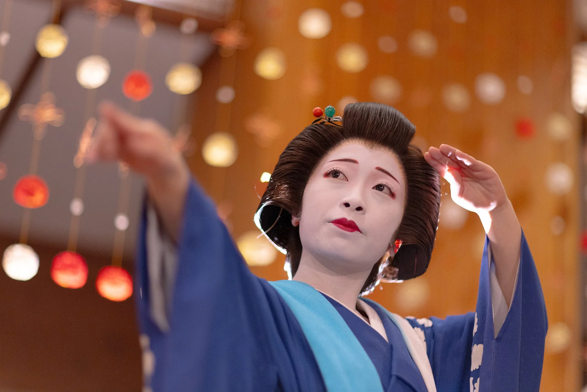 伝統的な白い化粧と赤い口紅、日本髪の女性が青い着物をまとい、指をさすようなポーズをとっています。背景には暖色系の玉ボケが広がり、舞台上でのパフォーマンスを思わせる一枚です。