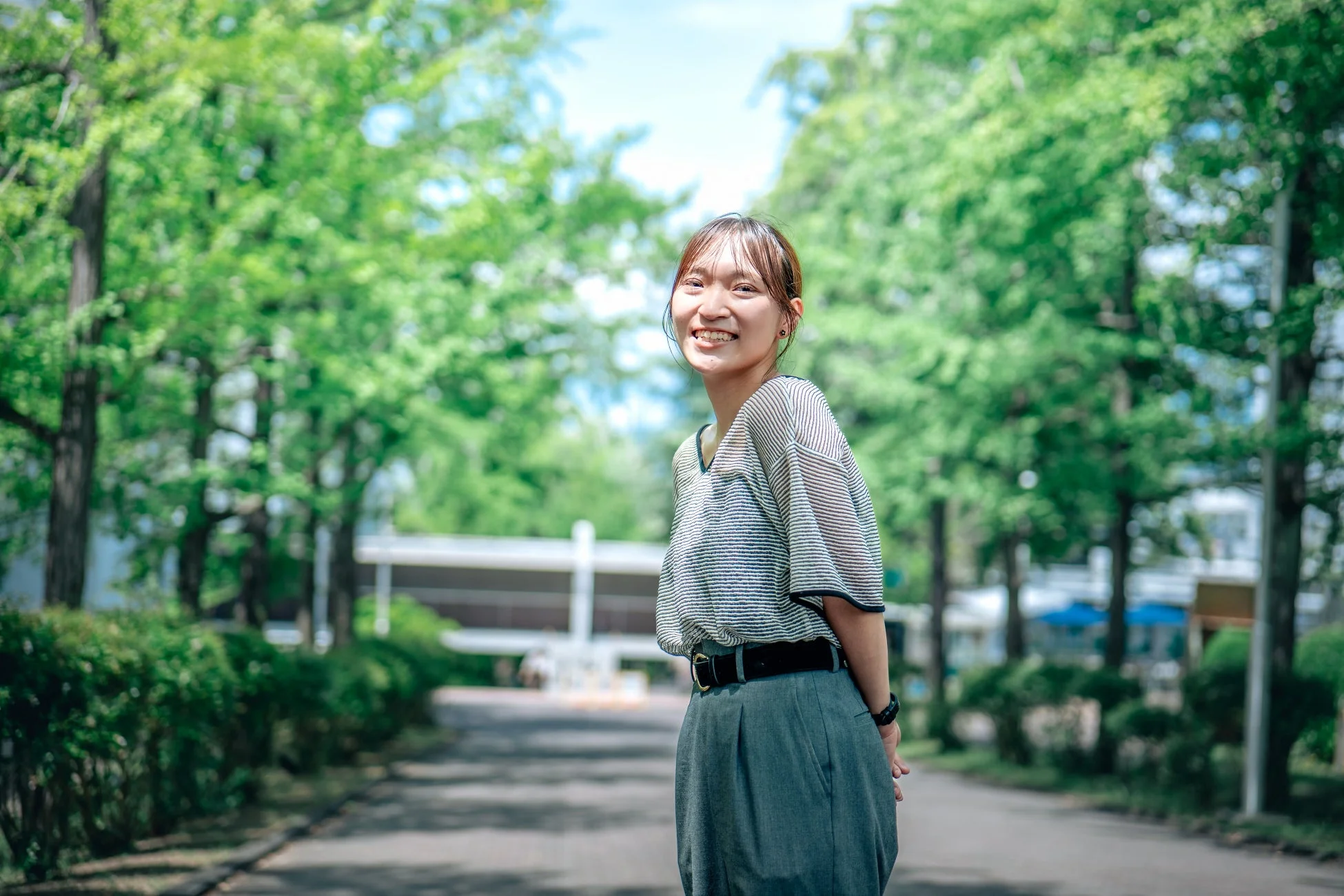 緑豊かな屋外で、カジュアルな服装の若い女性がカメラに向かって明るく微笑んでいます。