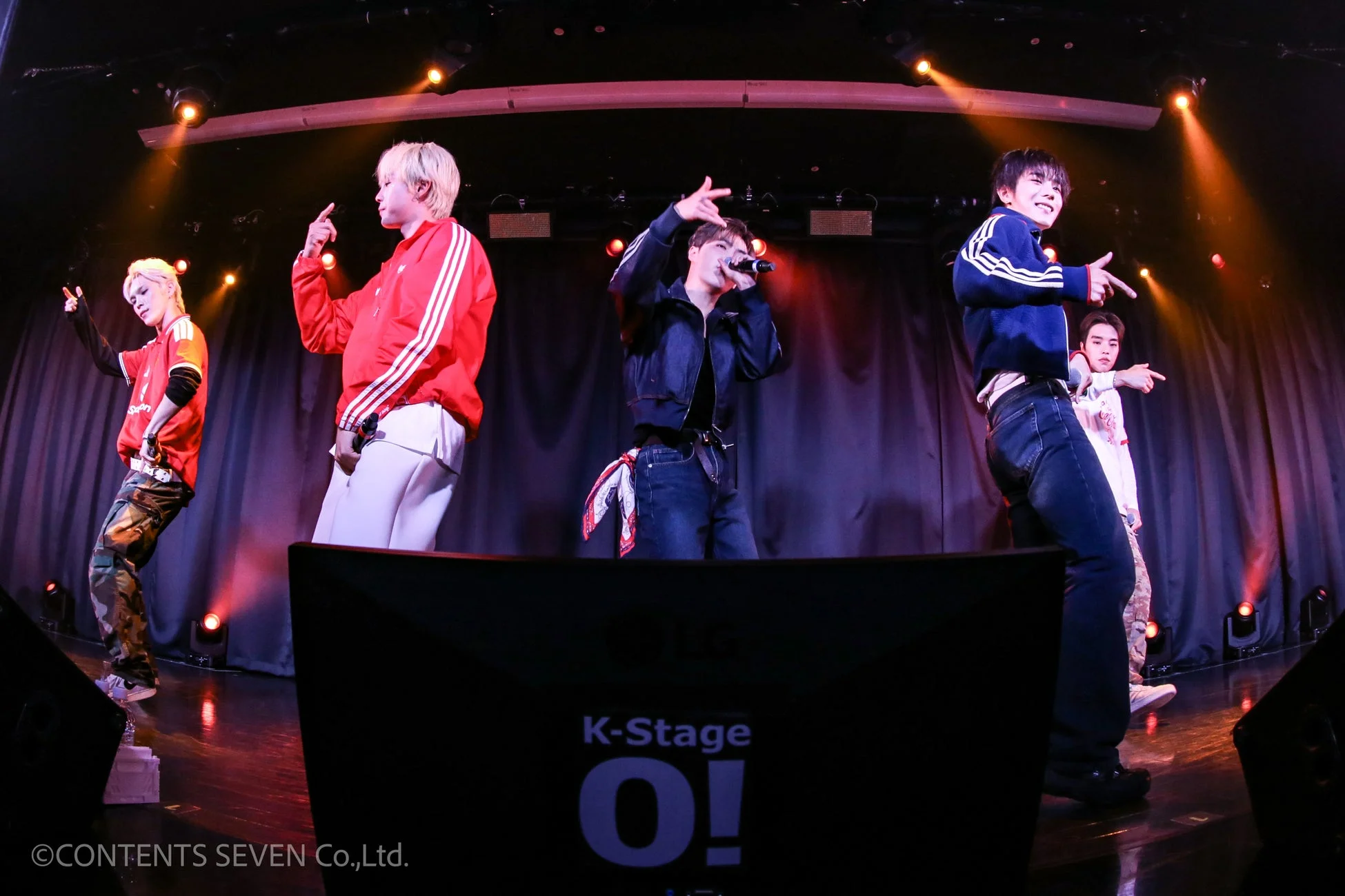 K-Stageのステージで、スポットライトを浴びながらパフォーマンスをする5人組の男性グループ。一人がマイクを持ち歌い、他のメンバーもエネルギッシュなポーズをとっている。
