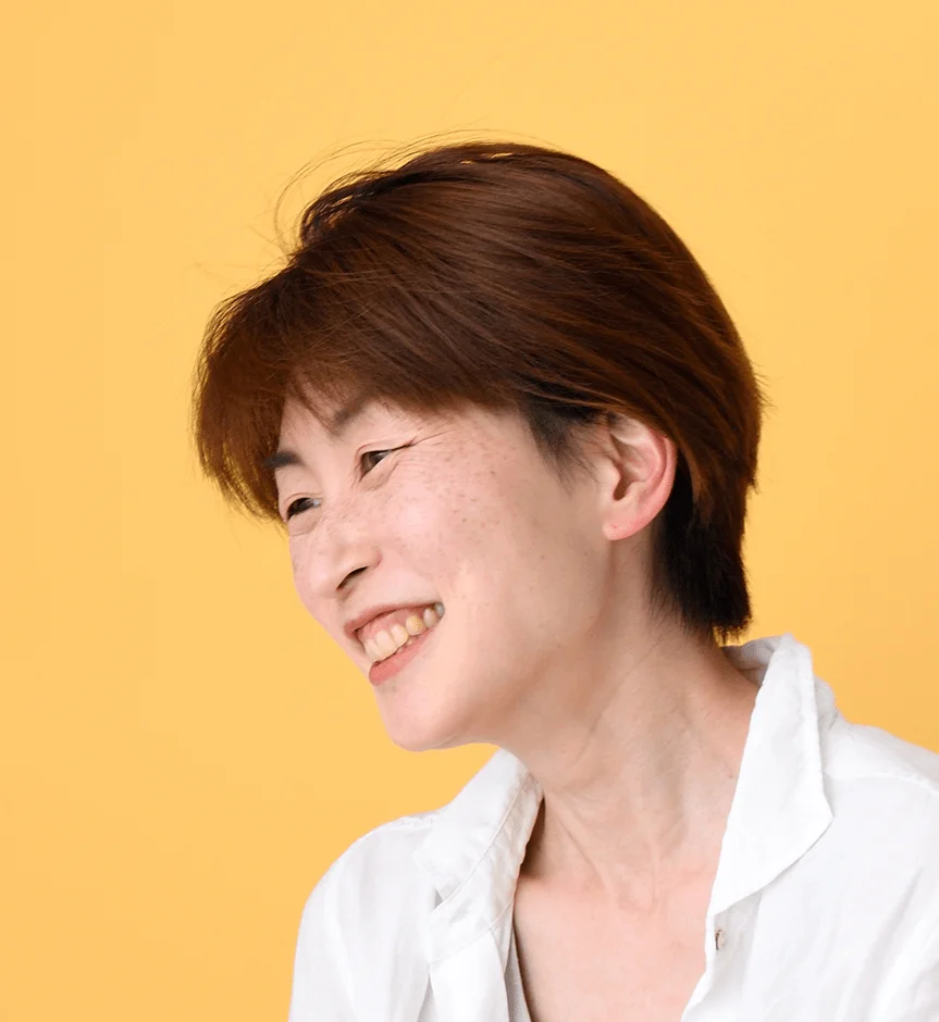 黄色い背景の前で、横向きに明るく笑うショートヘアの女性のポートレート。自然な笑顔が印象的です。