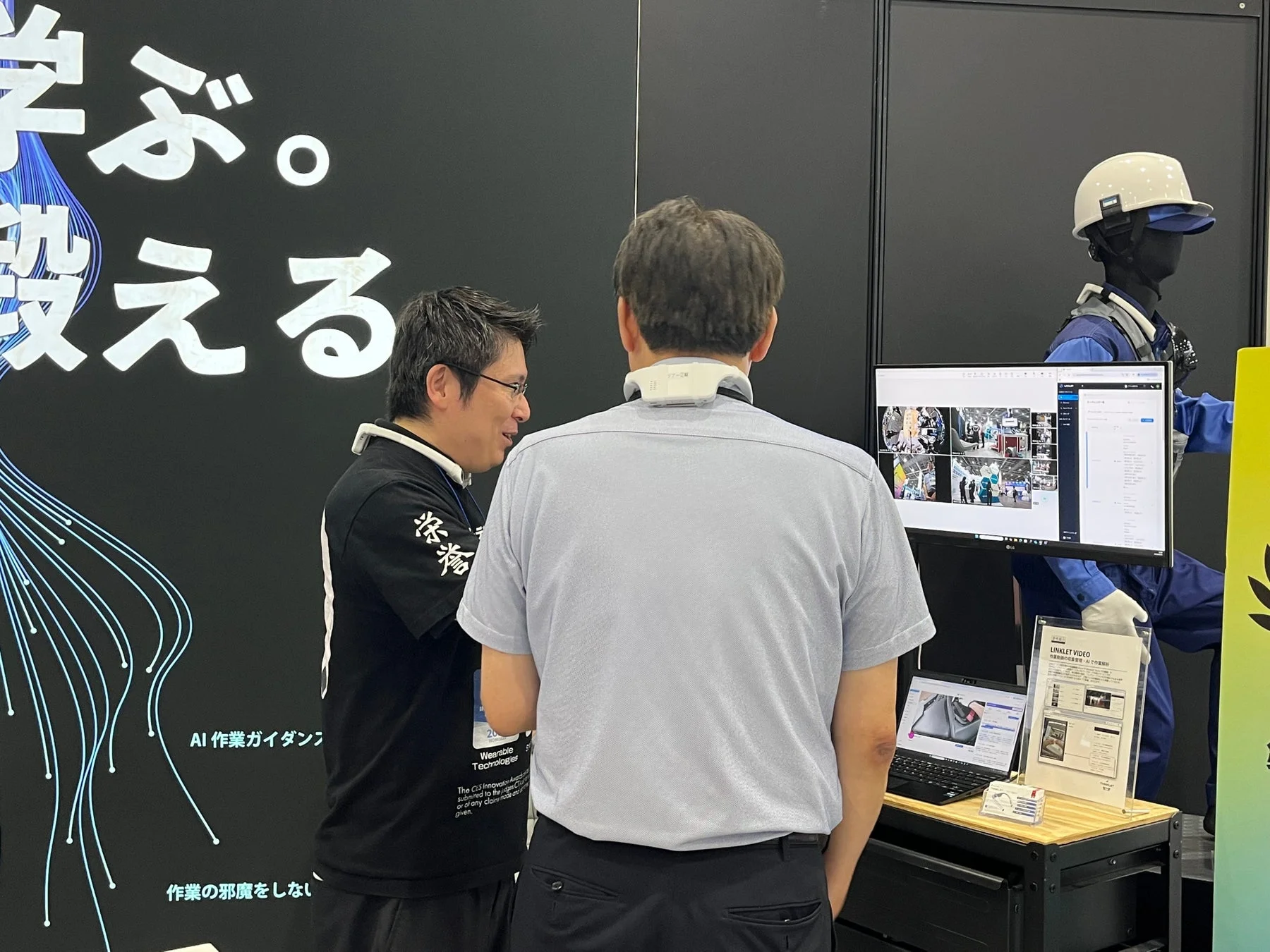 展示会ブースで、AI作業ガイダンスやウェアラブル技術について説明が行われている様子。一人の男性が首にデバイスを着用し、もう一人の男性が説明している。モニターには作業現場の映像が表示されている。