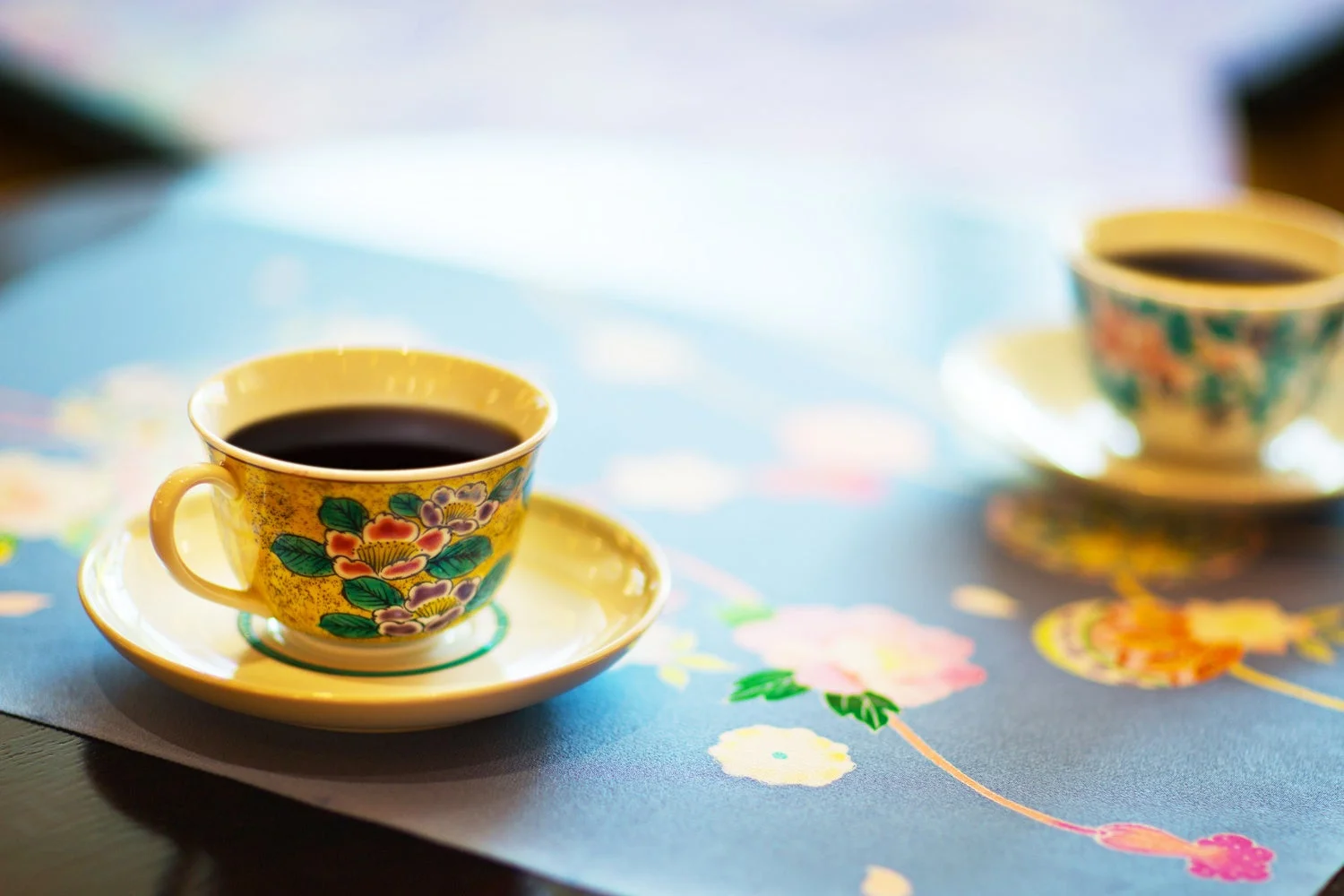 美しい花柄のカップ&ソーサーに入ったコーヒーが、花模様のランチョンマットの上に置かれています。手前のカップが鮮明で、優雅なひとときを感じさせます。
