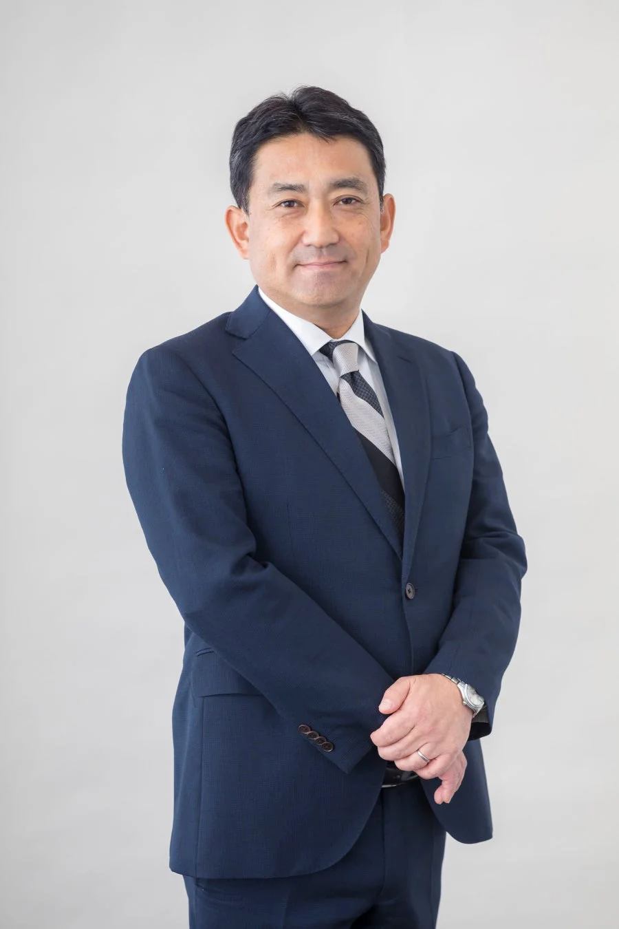 紺色のスーツを着た日本人男性が、白いシャツとストライプのネクタイを着用し、穏やかな笑顔でカメラを見つめている