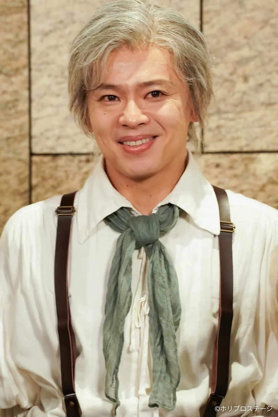 白髪の男性が、白いシャツに緑色のスカーフとサスペンダーを着用し、穏やかな笑顔をカメラに向けています。舞台衣装のような服装で、親しみやすい表情が印象的です。