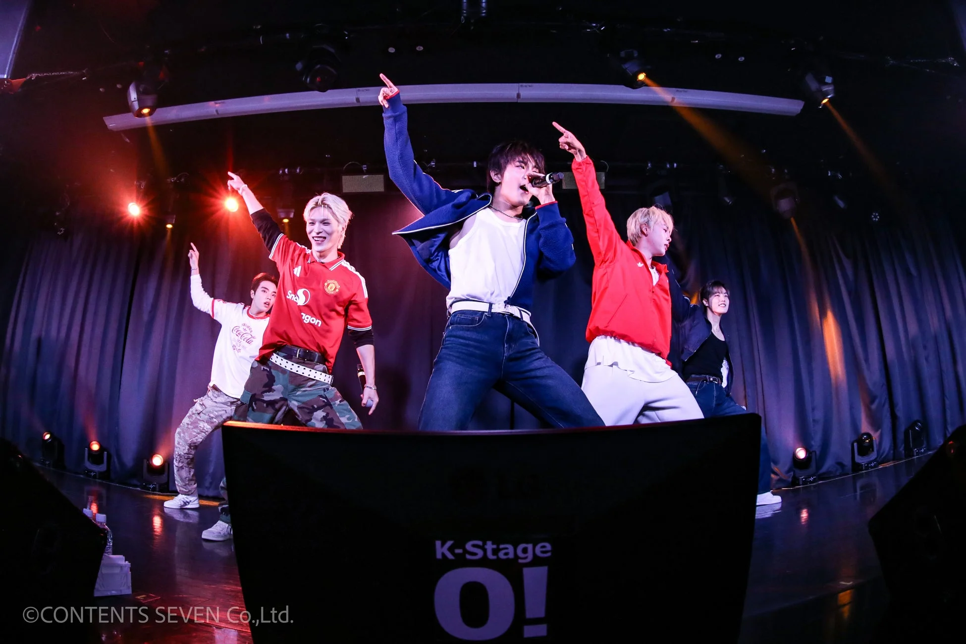 「K-Stage O!」のロゴがあるステージで、5人の男性グループがエネルギッシュなパフォーマンスを披露している。スポットライトが照らす中、メンバーが歌い、手を上げてポーズをとっている様子が捉えられている。