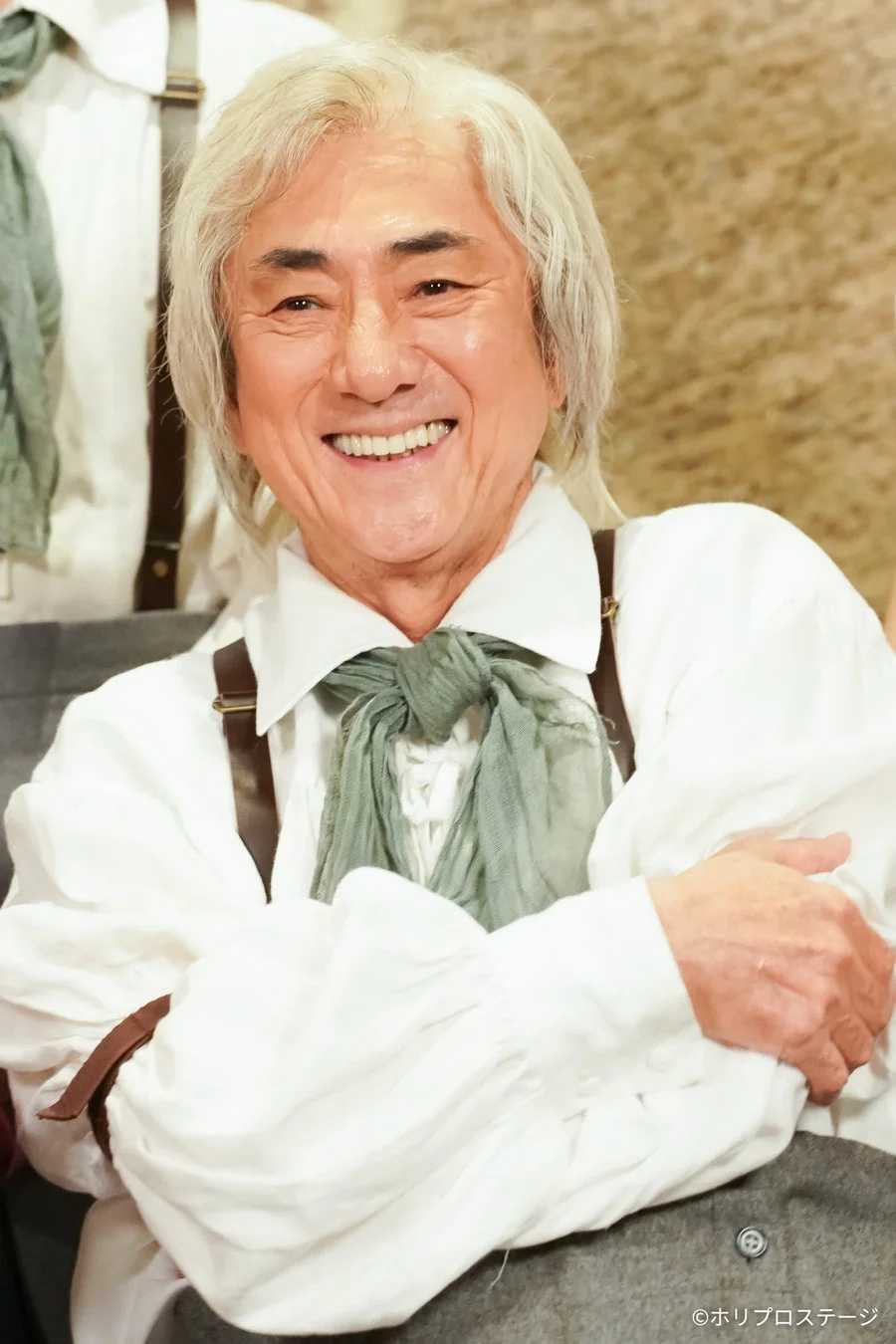 白髪の男性が白いフリルシャツに緑のスカーフとサスペンダーを着用し、腕を組んでにこやかに微笑んでいるポートレート。舞台衣装のような服装で、楽しそうな雰囲気が伝わる一枚です。